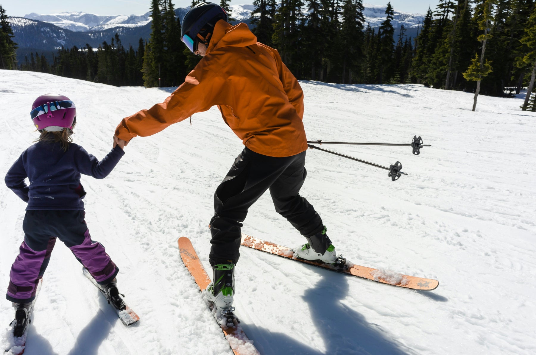 An adult holding a child’s hand as they ski down a snowy slope together.