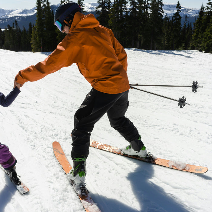 An adult holding a child’s hand as they ski down a snowy slope together.